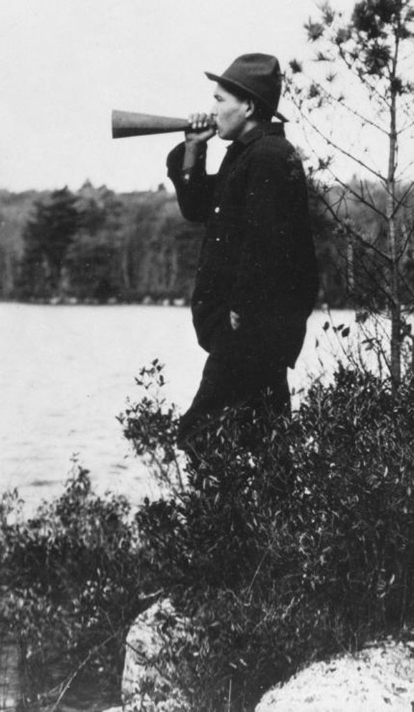 Old black and white picture of a man blowing on a horn while standing on the shores of a lake