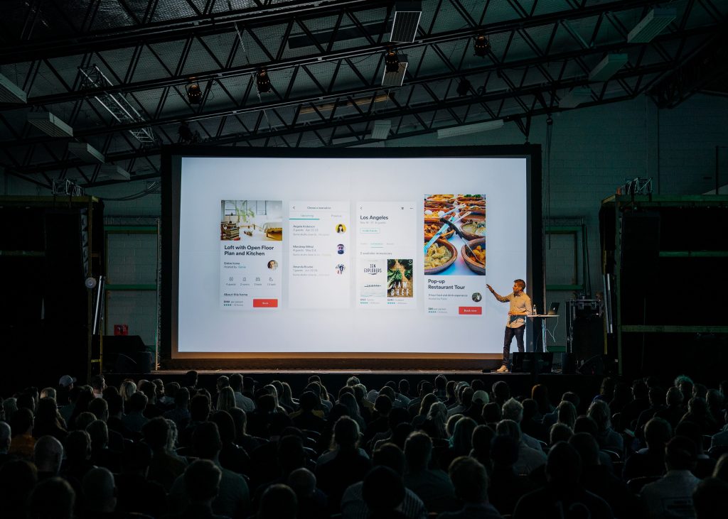 Man presenting on stage with a large screen behind him