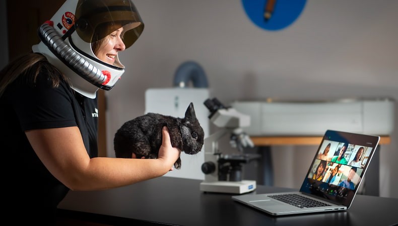 Woman in an astronaut helmet holds a cat in front of a laptop showing an online meeting