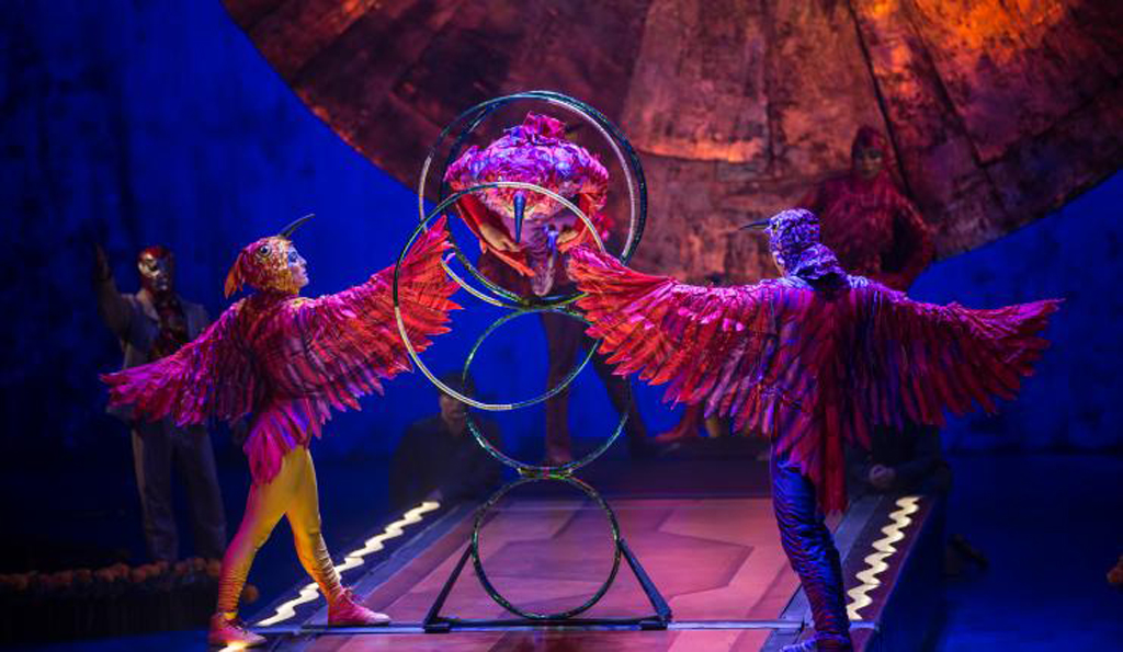 Two circus performers dressed as birds holding hoops that another performer is jumping through